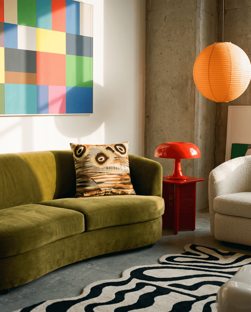 A modern living room scene featuring a curved olive green velvet sofa with a patterned throw pillow, a red side table with a red mushroom-shaped lamp, a concrete wall, and an orange paper lantern hanging light.