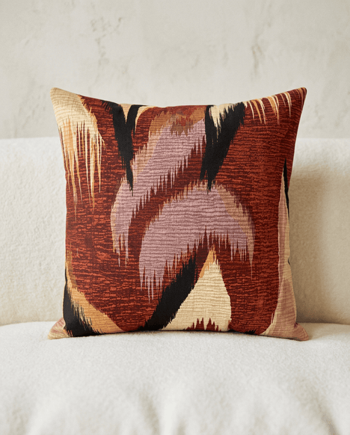 A square decorative throw pillow with an abstract ikat-style pattern in rust red, dusty pink, beige, and black, resting on a textured white sofa against a neutral wall.