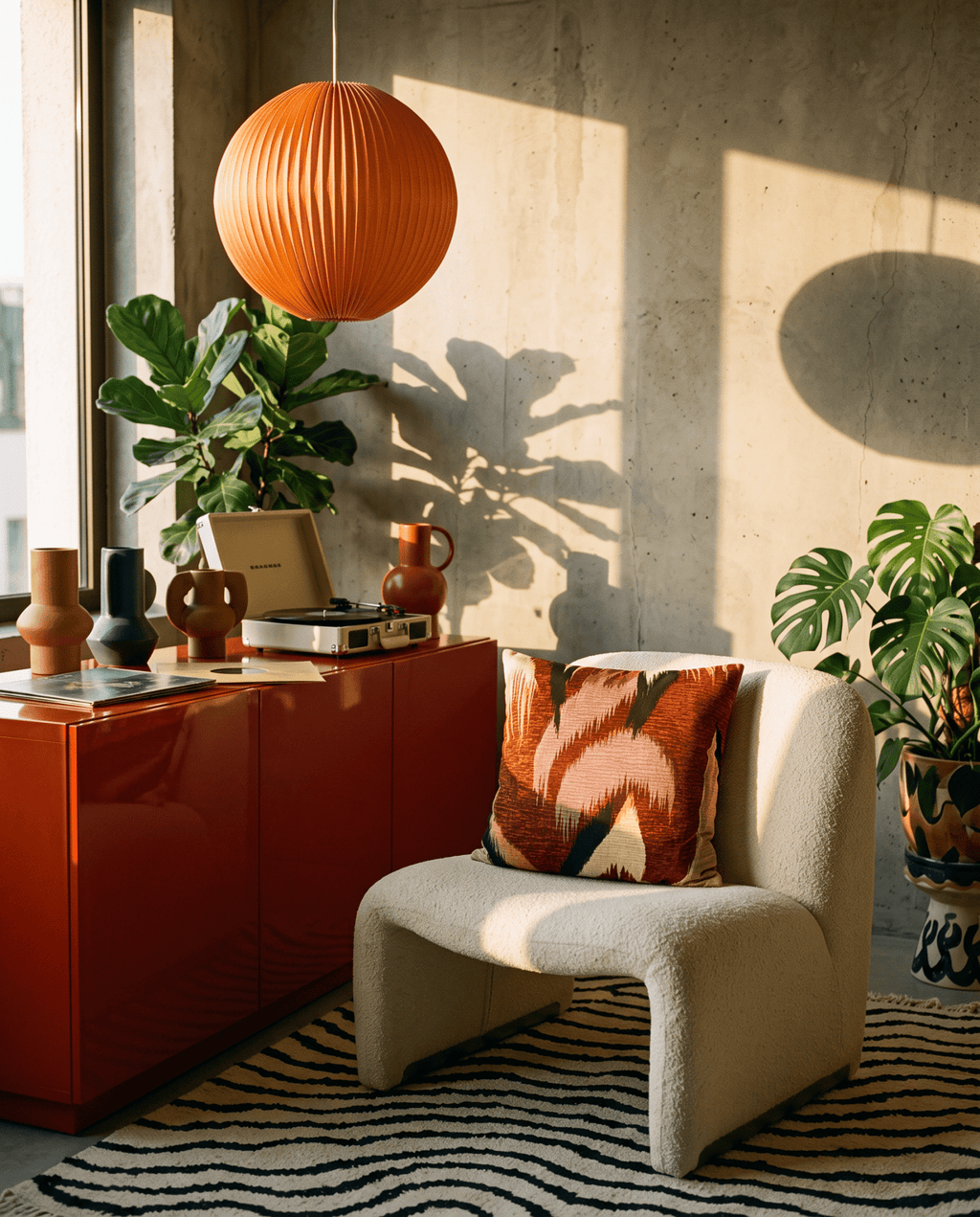 A modern living room corner featuring a cream bouclé armchair with a rust and orange patterned throw pillow, set next to a glossy red sideboard with decorative vases and plants. A large orange pleated paper lantern hangs from the ceiling, casting a shadow on the concrete wall.
