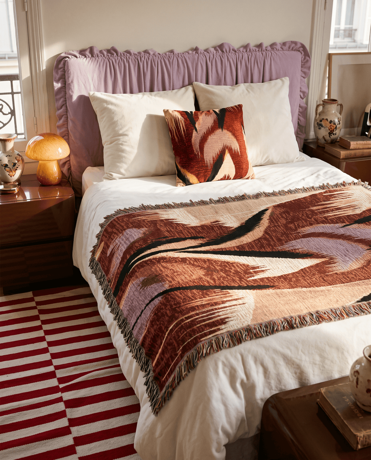A styled bedroom scene showing a bed with a white duvet, a mauve ruffled headboard, a patterned throw blanket in rust, brown, and cream, cream pillows, and a red-and-white striped rug on the floor.