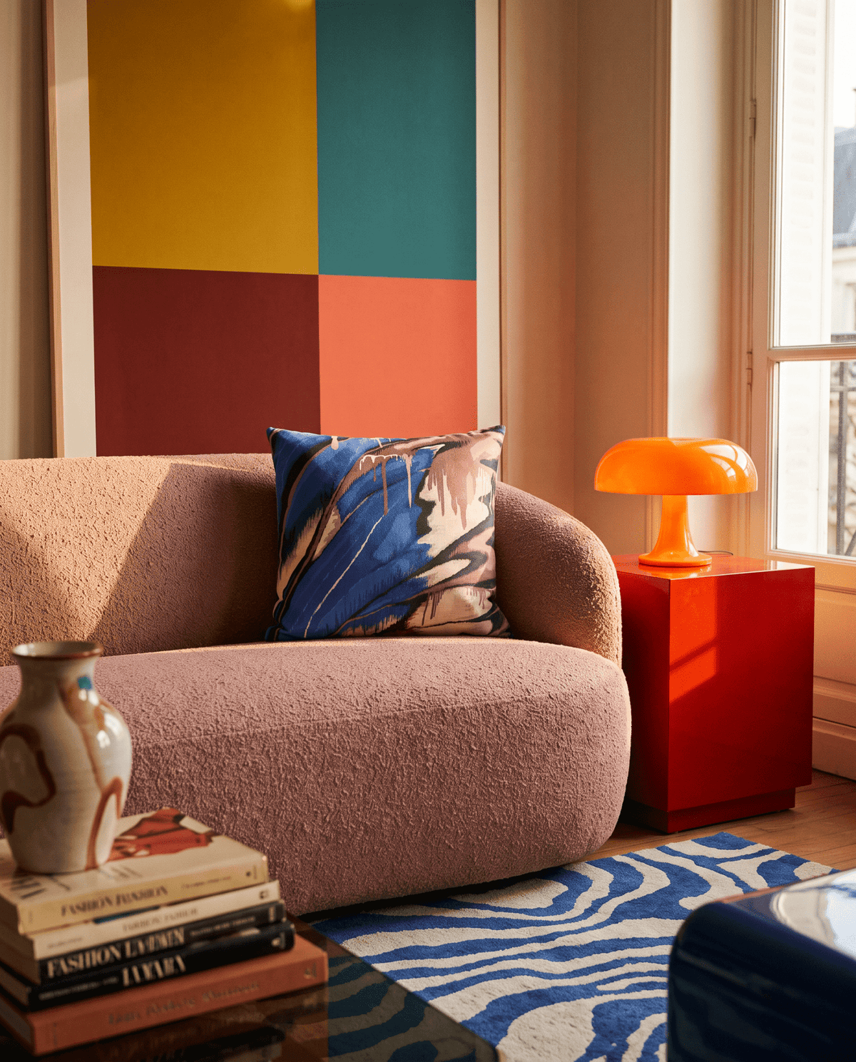 A styled interior scene featuring a blush pink boucle sofa with a blue and beige botanical patterned accent pillow, set in a room with a colorful geometric wall art piece, a red side table with an orange lamp, and a blue patterned rug.