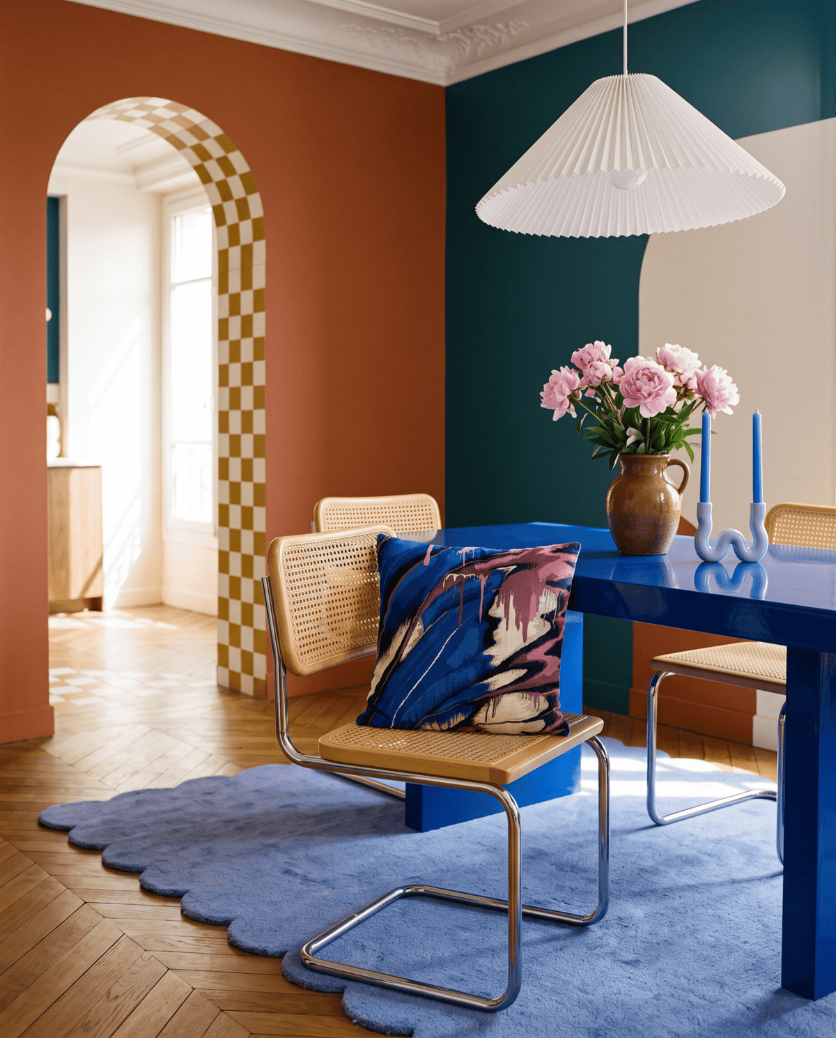 A modern dining room corner featuring a vibrant blue table, woven cane chairs, a blue scalloped rug, and a colorful abstract-patterned throw pillow. The room has bold color-blocked walls in terracotta, deep teal, and cream, with a white pleated pendant light hanging above.