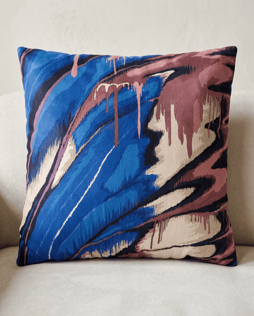 A square throw pillow featuring an abstract paint-drip pattern in vibrant blue, dusty rose, cream, and black, resting on a light beige upholstered chair.