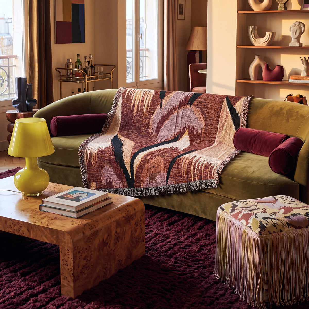 A styled living room scene featuring a green velvet sofa draped with a large woven throw blanket in earthy pink, brown, and black tones. A matching patterned fringed ottoman sits in the foreground beside a burl wood coffee table and a bright yellow lamp, all set on a deep burgundy shag rug.
