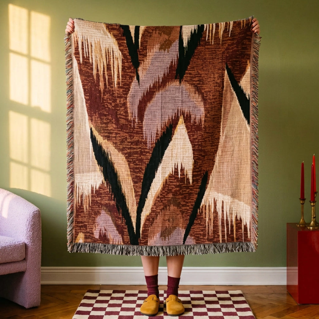 A person stands in a room holding up a large woven throw blanket with a geometric pattern in brown, beige, black, and muted pink. The room has olive green walls, a checkered rug, and furniture including a lavender armchair and a red side table with red candles.