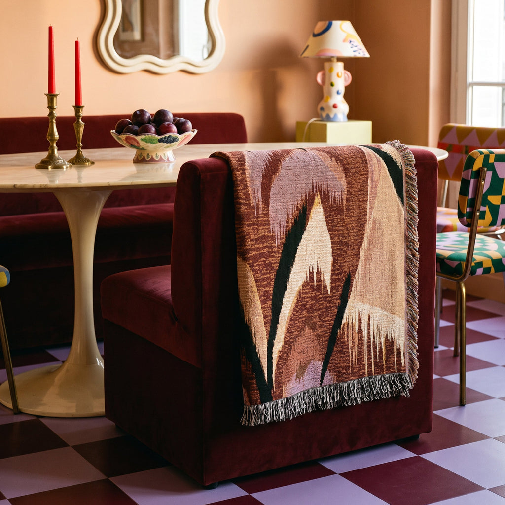A woven throw blanket with a geometric mountain pattern in brown, beige, and black hangs over the back of a deep burgundy velvet armchair in a retro-styled dining room.