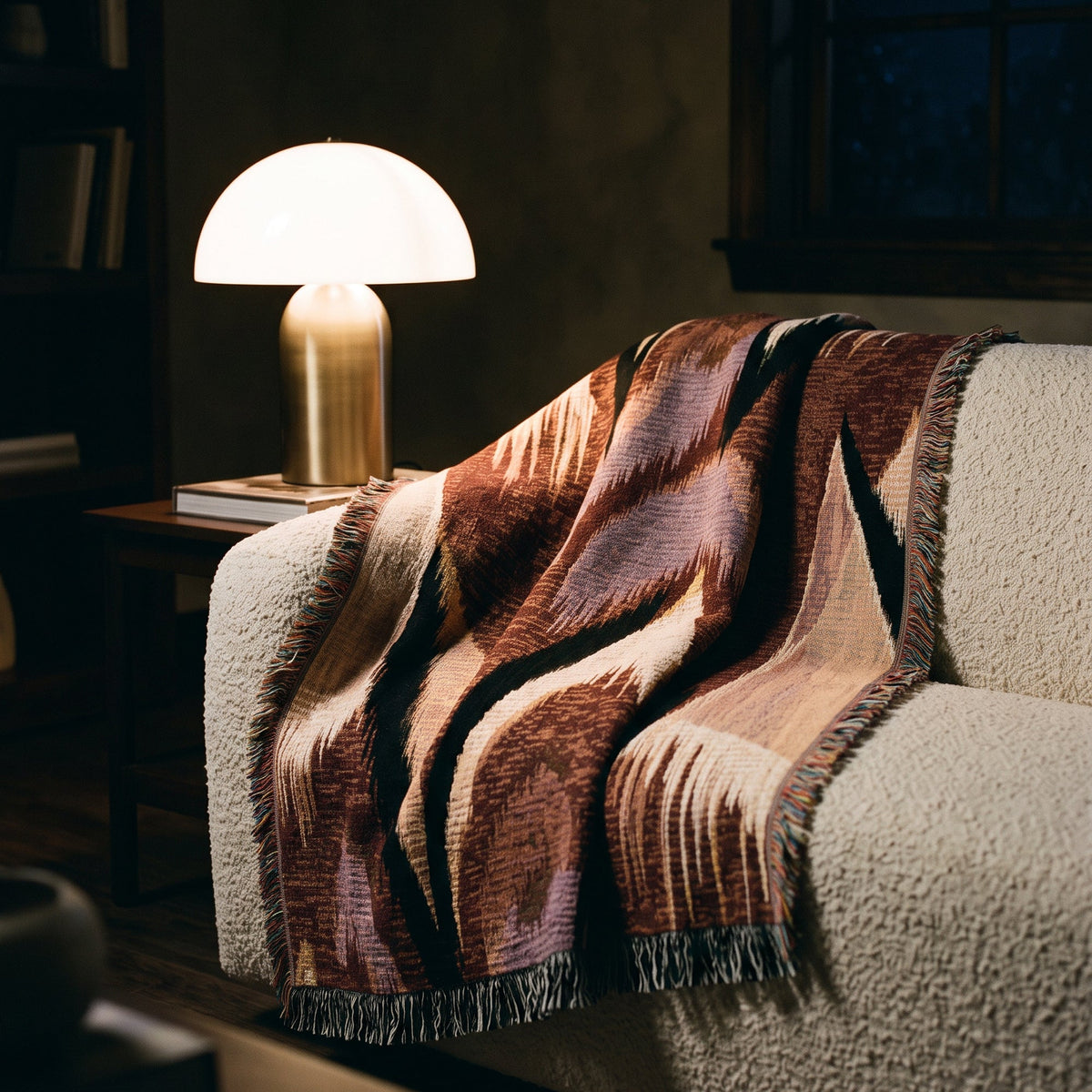 A patterned woven throw blanket draped over a textured cream sofa in a warmly lit living room, next to a glowing table lamp.