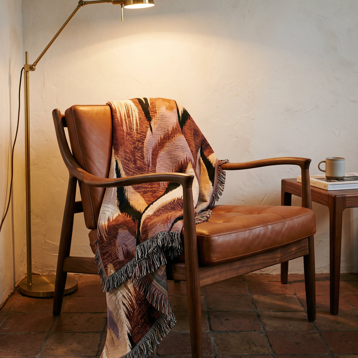 A mid-century modern wooden armchair with brown leather upholstery, styled with a patterned brown and beige fringed throw blanket. The chair is set in a cozy room with a warm brass floor lamp, a small wooden side table with a mug, and a textured off-white wall.
