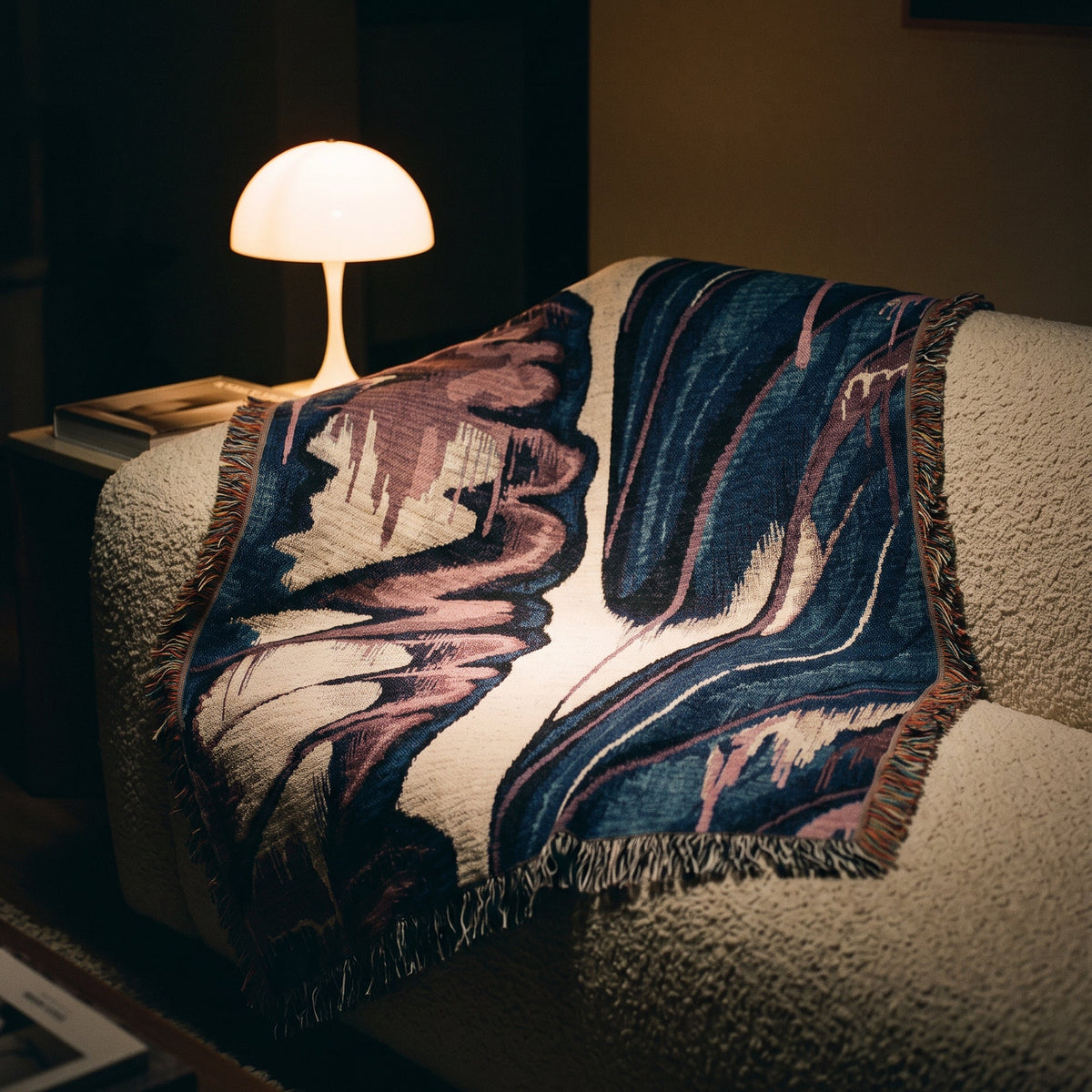 A woven throw blanket with an abstract marbled pattern in navy, blue, cream, and dusty pink, draped over a textured beige armchair in a warmly lit room next to a glowing mushroom-shaped table lamp.