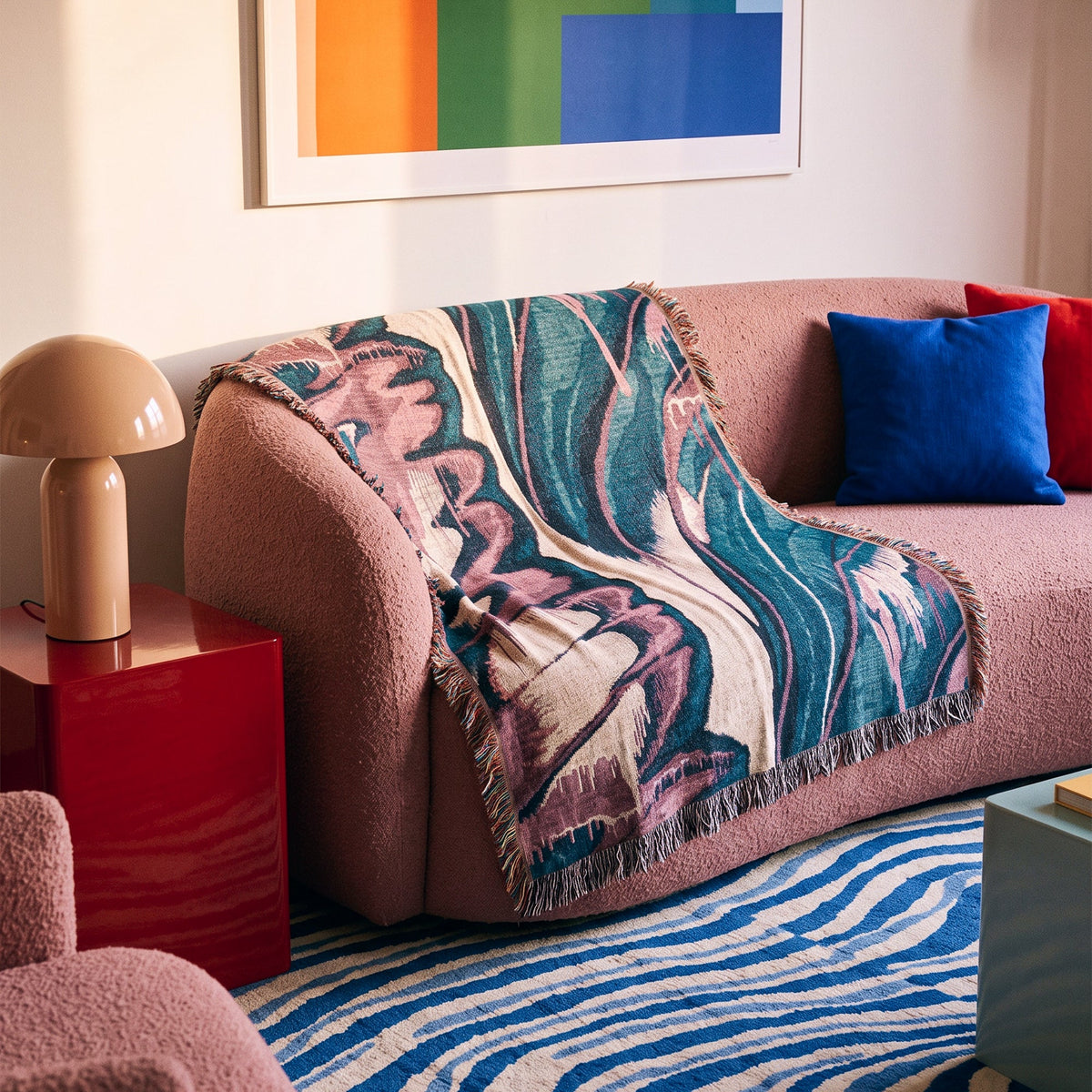 A textured mauve-pink bouclé sofa in a modern living room setting, styled with a colorful abstract patterned throw blanket, a blue pillow, and a red side table.