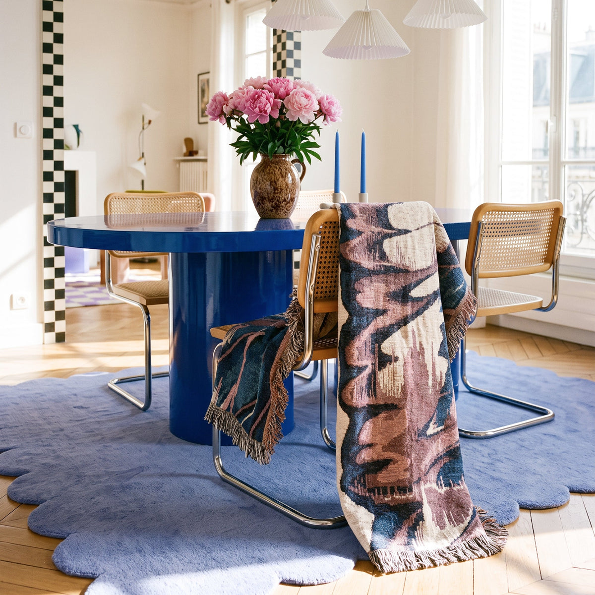 A styled dining room scene featuring a round blue table, woven cane chairs, a large scalloped blue rug, and a patterned fringed throw blanket draped over the chairs. A vase of pink flowers sits on the table.