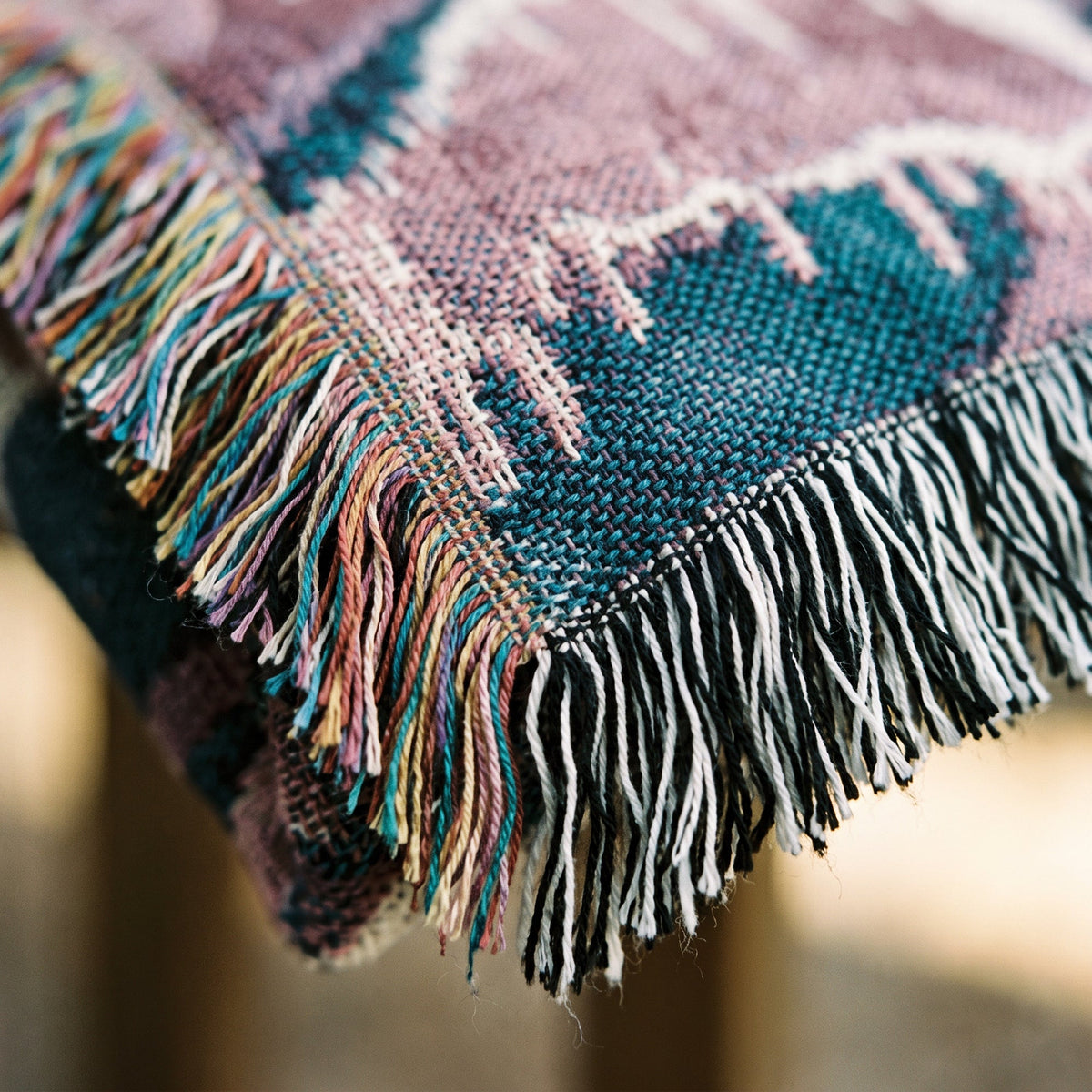 Close-up of a woven textile with multicolored fringe, showing a dusty pink and teal patterned fabric with black and white fringed edges.