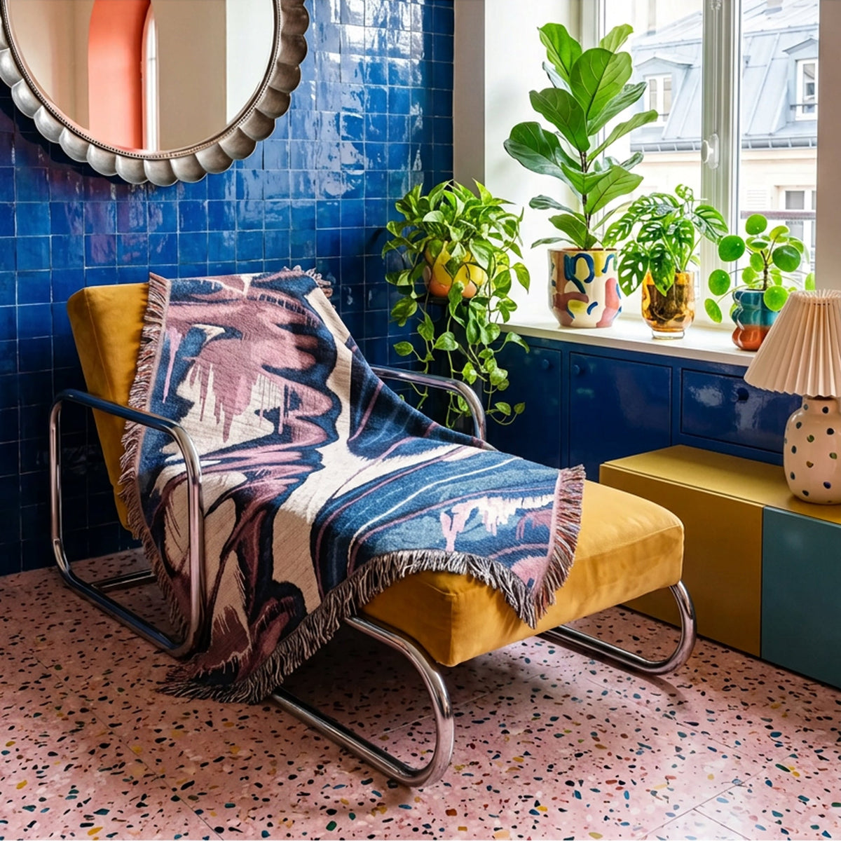 A mustard yellow velvet lounge chair with a chrome frame, styled with a multicolor abstract patterned throw blanket. The chair is set in a vibrant room with a blue tiled wall, a window with potted green plants, and a pink speckled terrazzo floor.