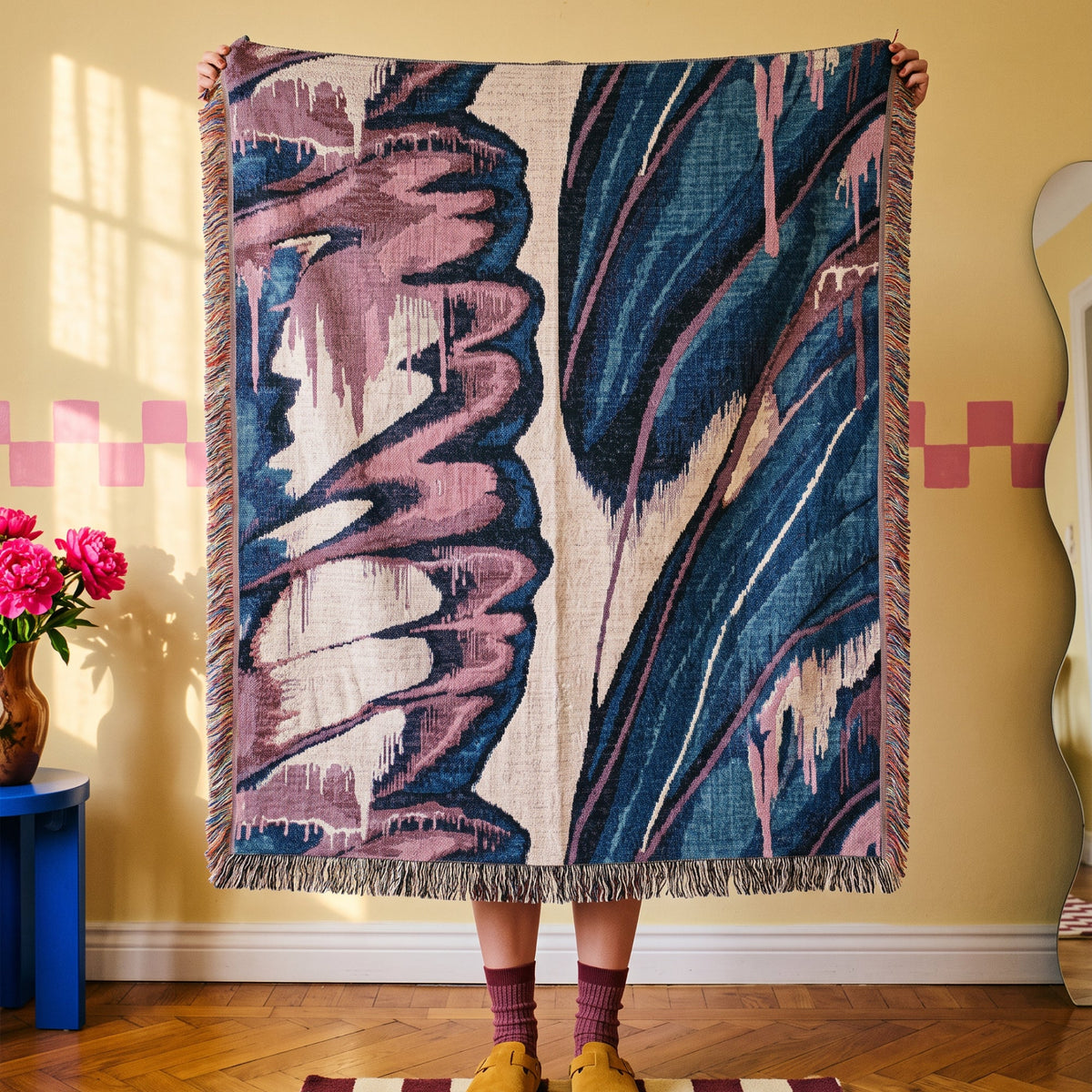 A person holds up a large woven throw blanket with fringe on all sides. The blanket features an abstract ikat-style pattern in navy blue, dusty pink, and cream.