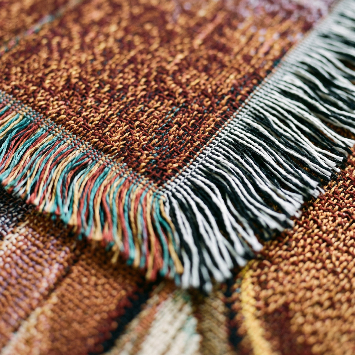 Close-up of a woven textile with a multicolor fringe border in black, white, teal, and rust tones over a brown and tan patterned background.