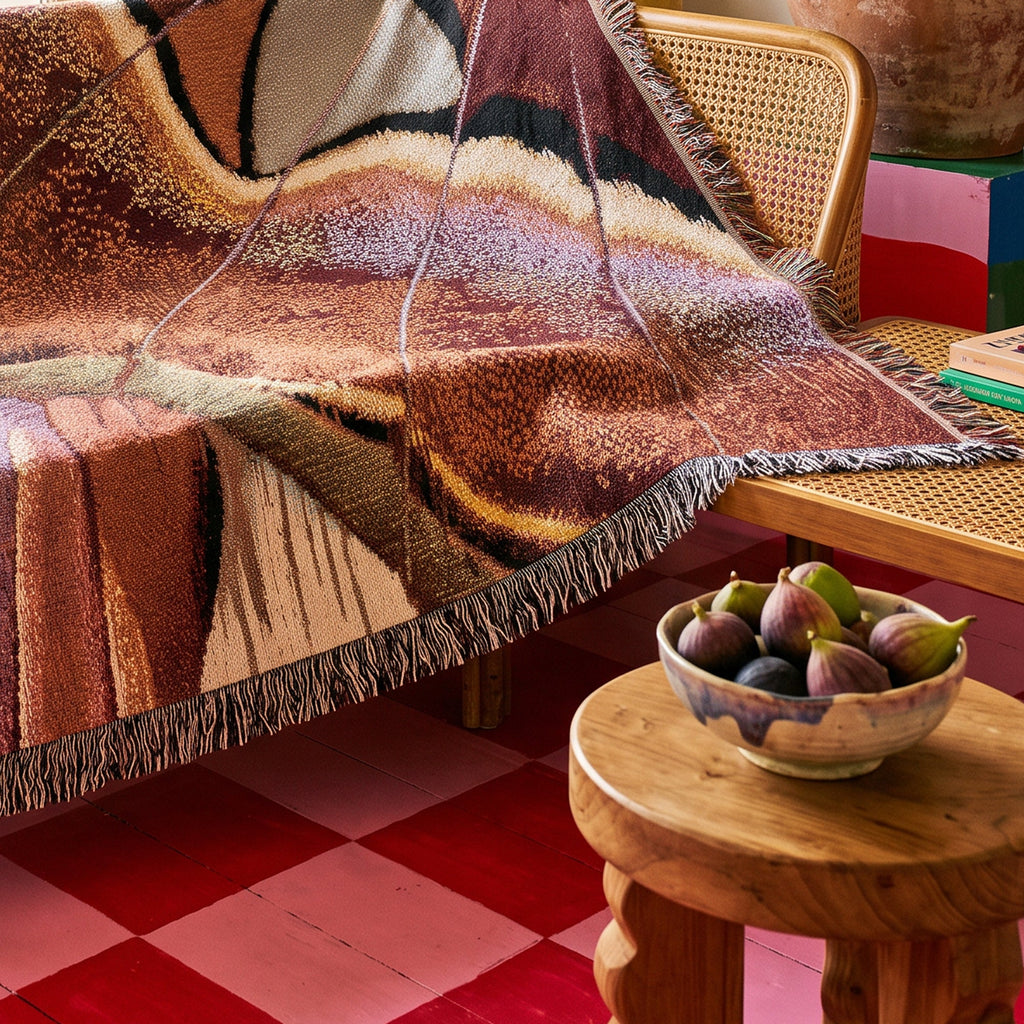 A woven throw blanket with a multicolor abstract pattern in earthy tones of brown, beige, and purple, draped over a cane-backed daybed in a styled room with a wooden stool and a bowl of figs.