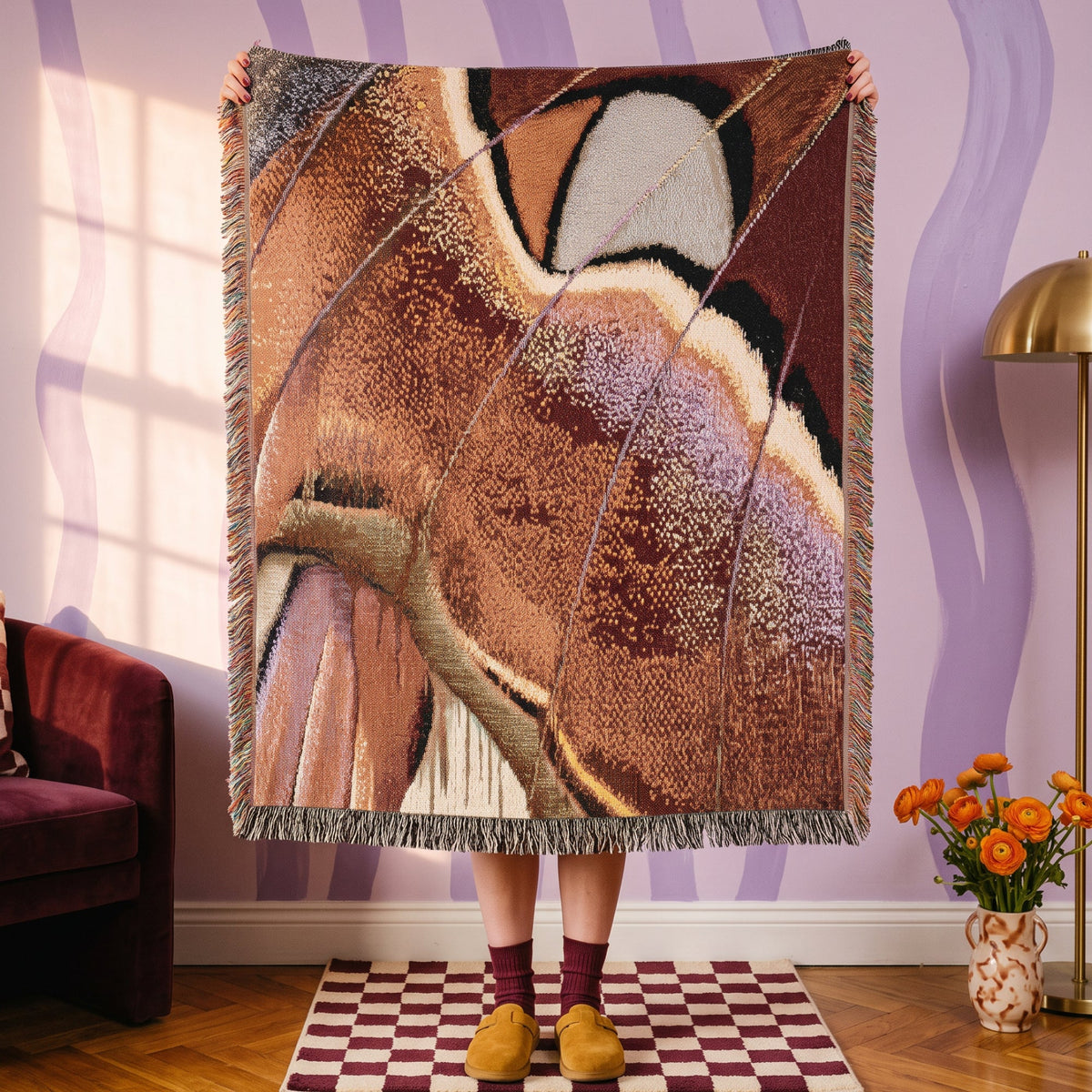 A person holds up a large woven throw blanket with fringe in a styled room. The blanket features an abstract, organic pattern in earthy tones of brown, beige, cream, and muted purple.
