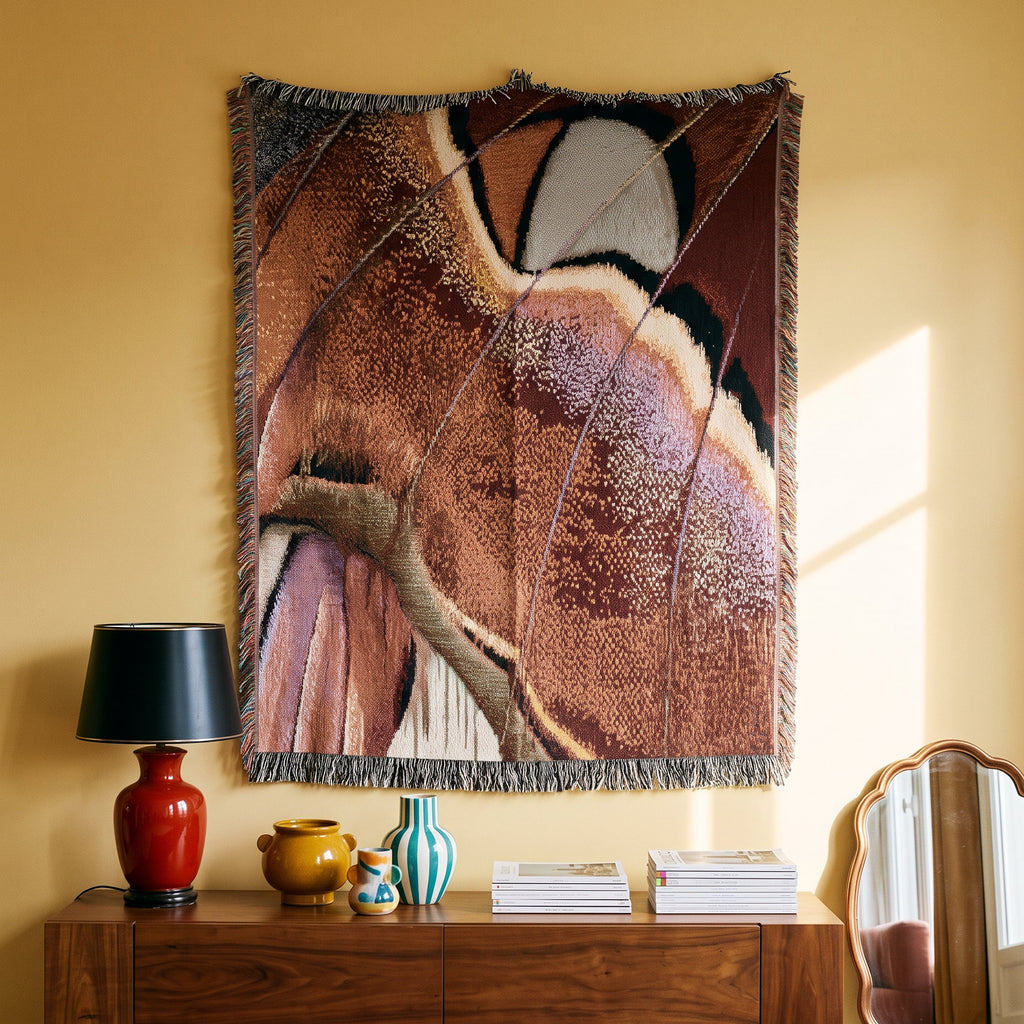 A large woven wall tapestry with abstract organic patterns in brown, beige, cream, and muted purple tones, hanging on a warm yellow wall above a wooden console table with a red lamp and decorative vases.